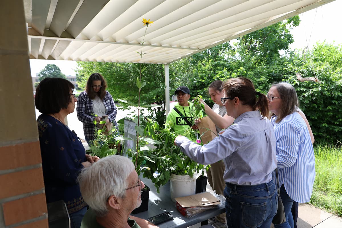 Nadia Navarrete-Tindall stands beside Native Plant Academy participants gathered around a table of potted native plants during a plant giveaway.