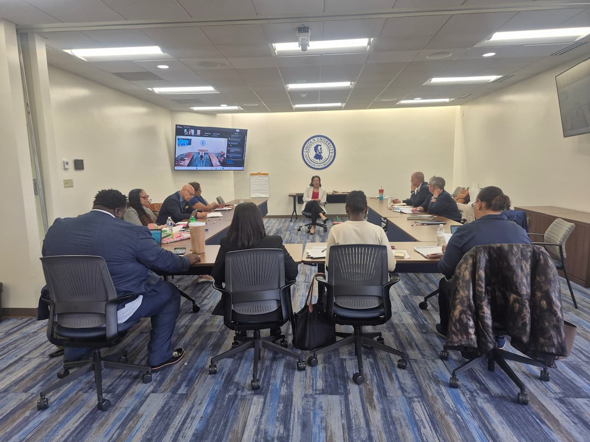 A group of people sit in a conference room at a table and chairs. 