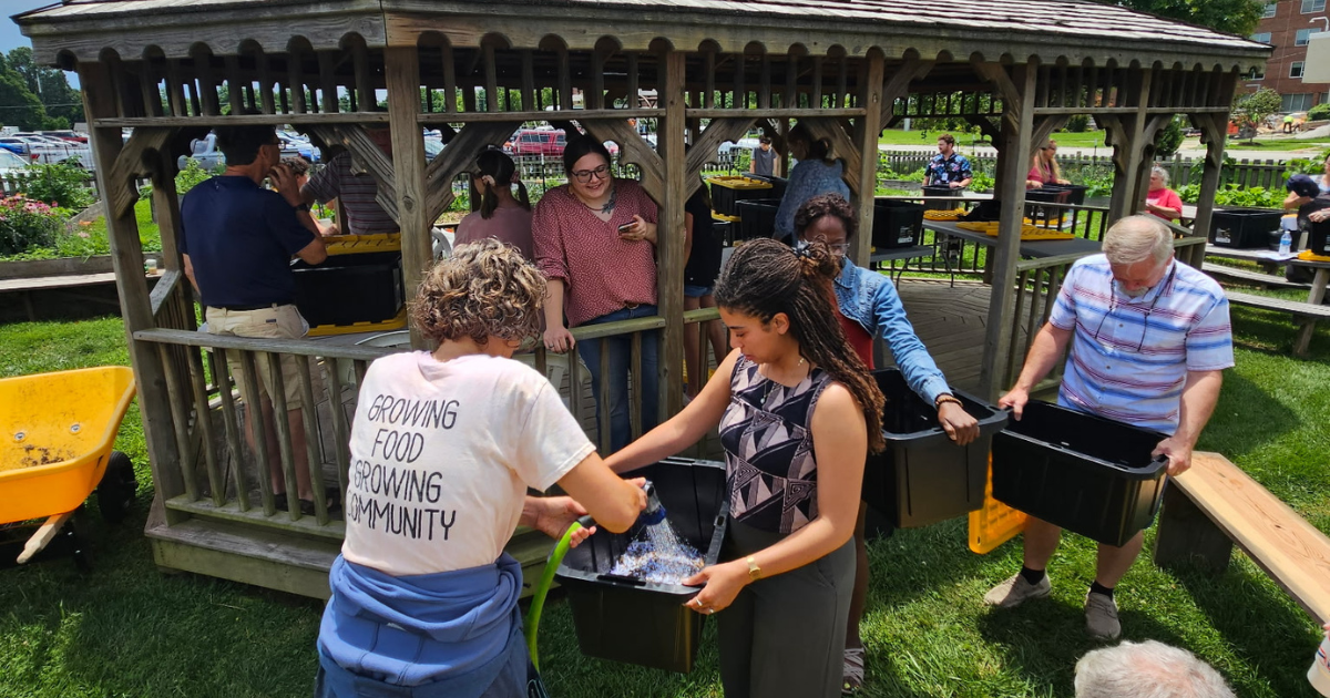 A group of people holding black containers with yellow lids gathered around a wooden gazebo in a grassy outdoor area, engaging in a gardening activity.