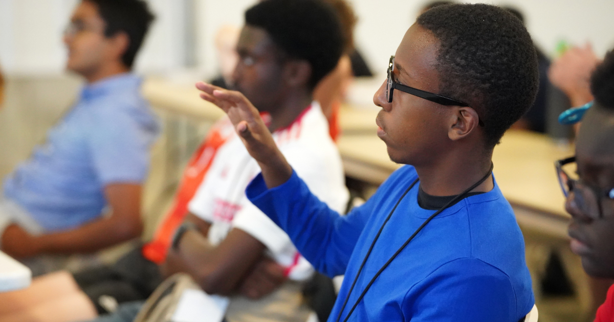 A student in a blue shirt raises his hand while seated in a classroom.