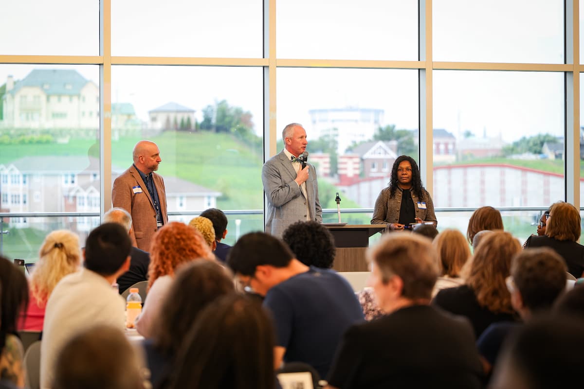 Three presenters speak to a large group of people sitting at round tables.