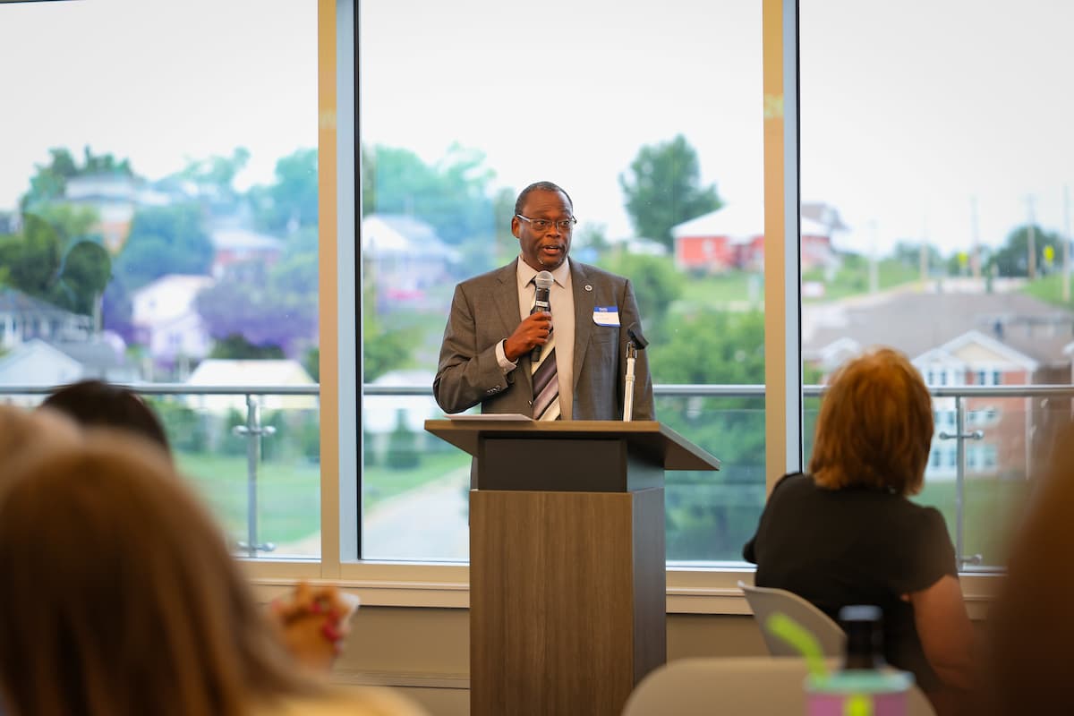A male presenter speaks to a large group of people sitting at round tables.