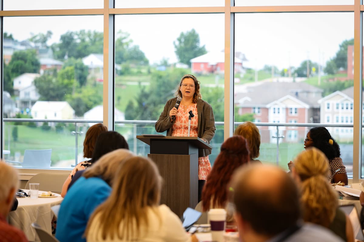 A female presenter speaks to a large group of people sitting at round tables.