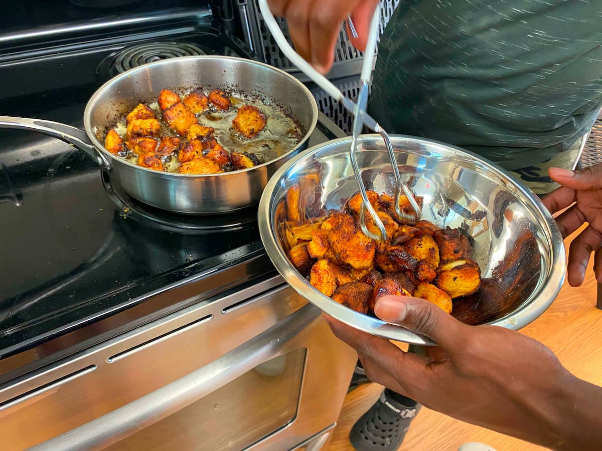 Vincent Osei uses tongs to transfer fried plantain pieces from a skillet into a metal bowl.