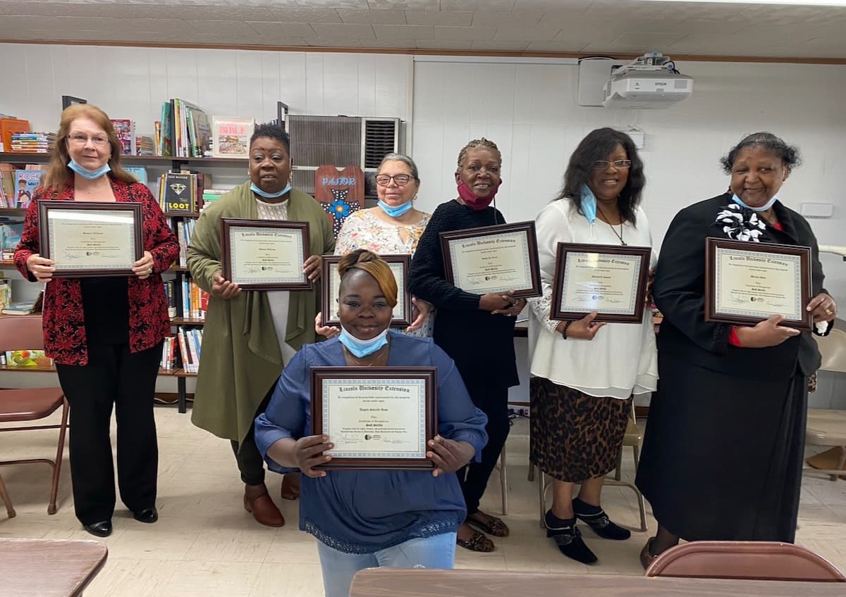 Seven women holding framed certificates in a library setting.  