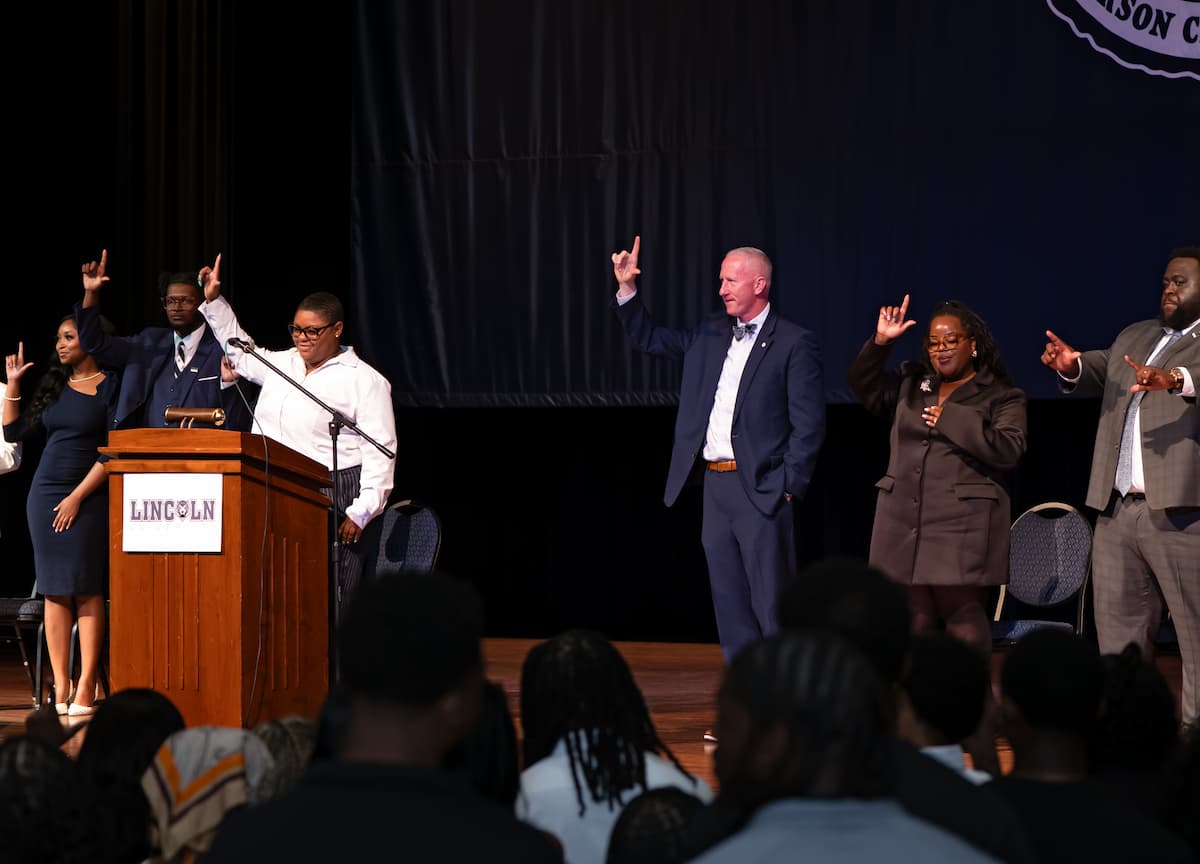 A group of people in business clothing stand on a stage while making an L with their hands.