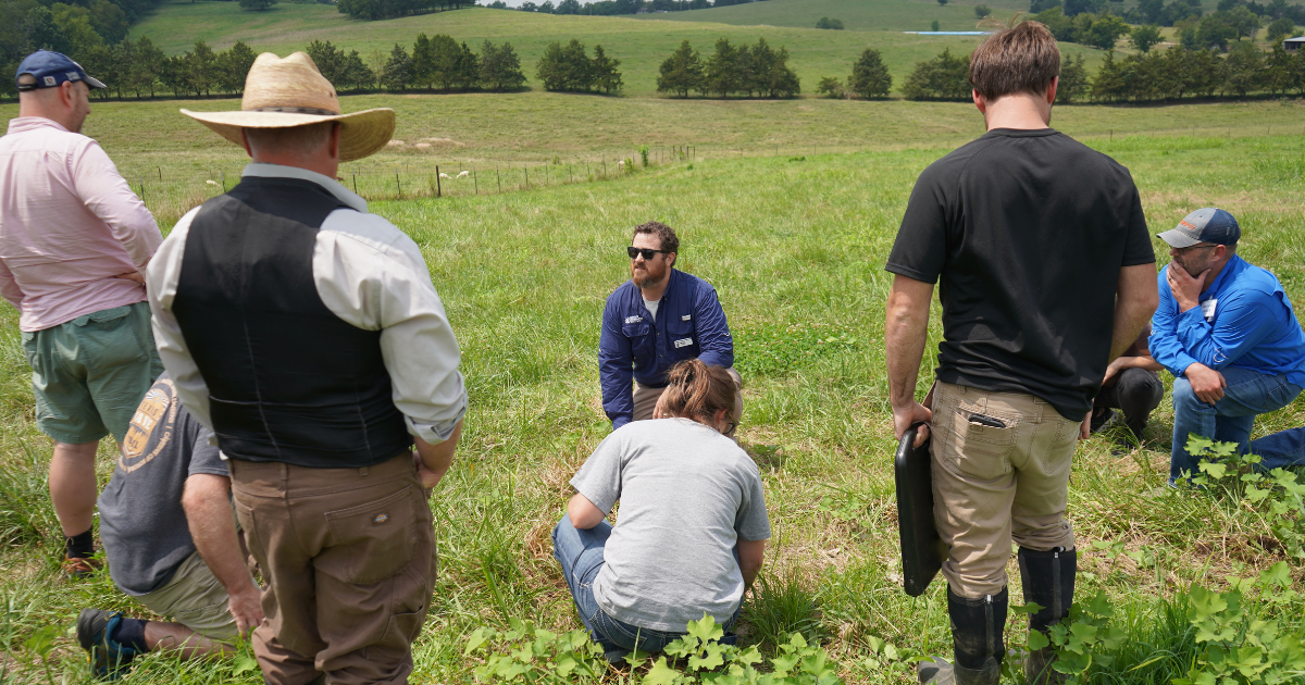 A group of seven people gathered in a grassy field. Two people in the middle of the group ares bent down touching the grass.
