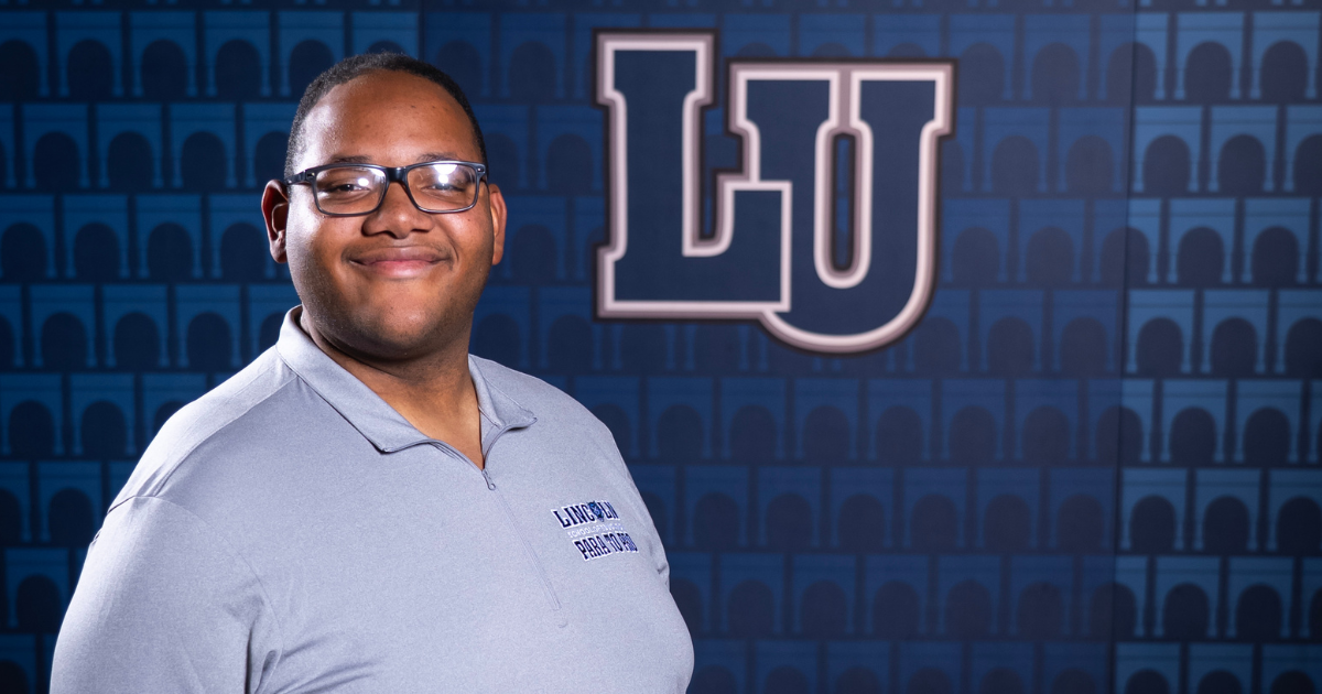 A young man with glasses in a blue shirt stands and smiles with a Lincoln University branded wall behind him.