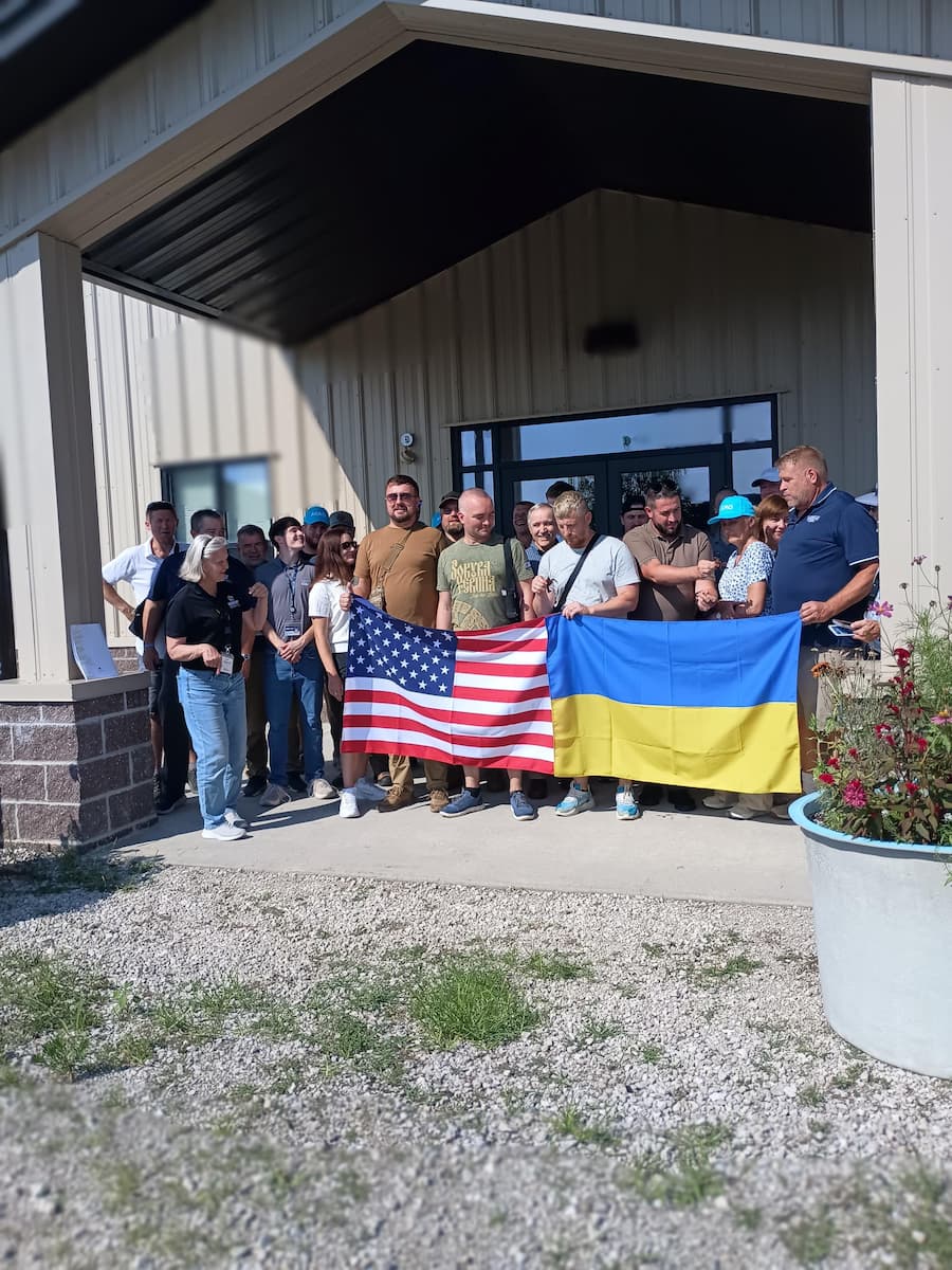 A group of men and women standing outside a building with Ukrainian and American flags in front of them.