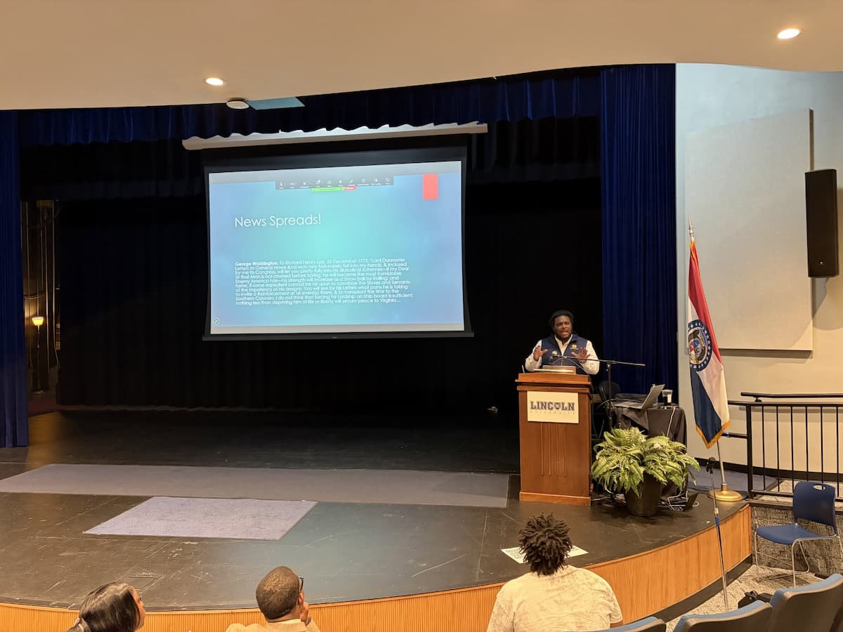 A man stands at a podium while giving a speech and a big presentation screen displays his work.