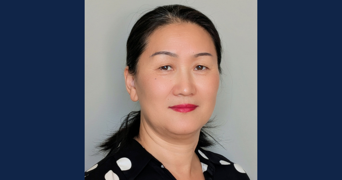 Photograph of a middle-aged Asian woman with short black hair wearing a dark blazer and light-colored top, set against a plain purple background.