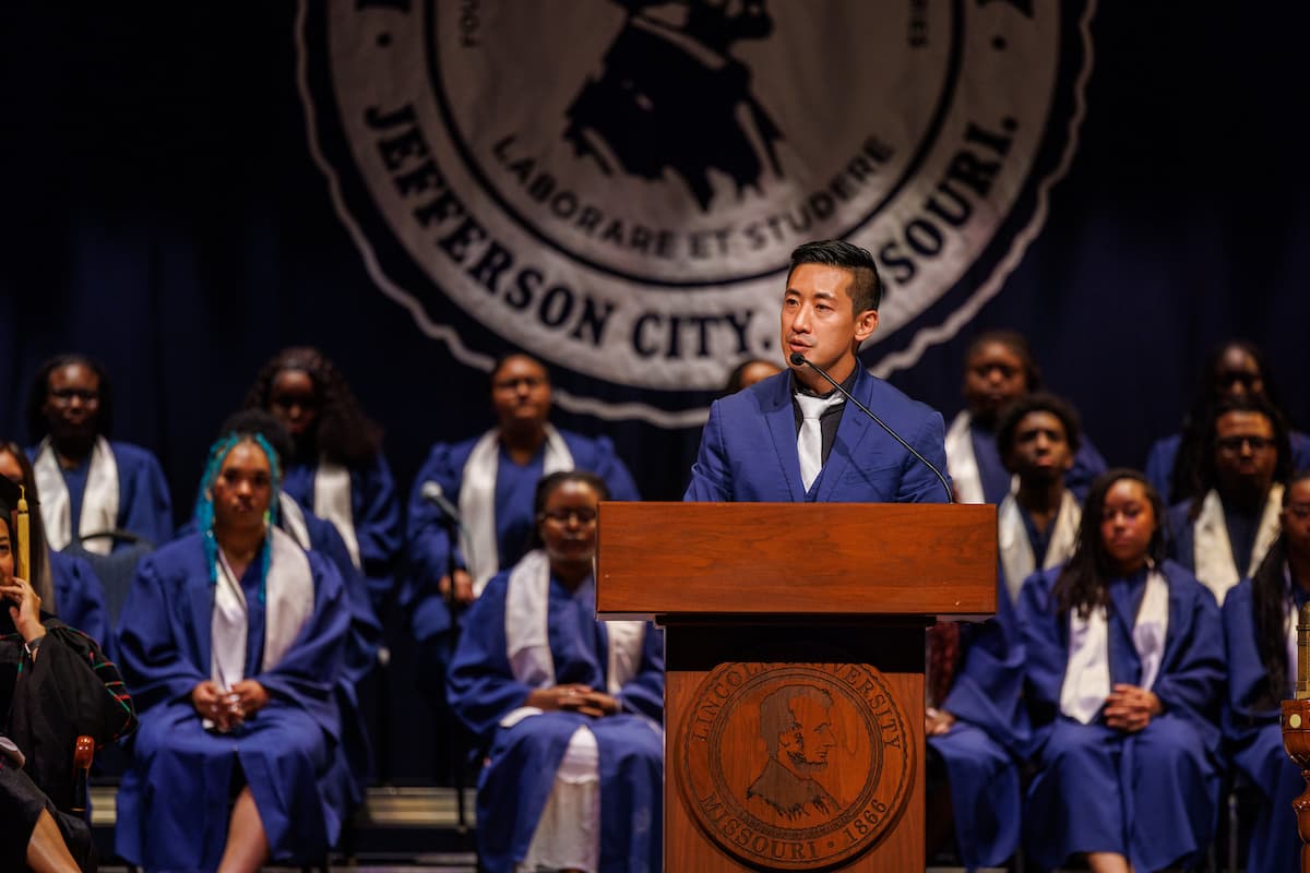 A man in a suit stands behind a podium while giving a speech on a stage.