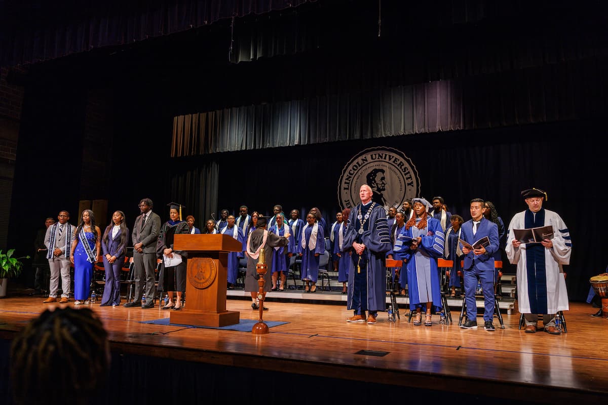 A group of people dressed in academic regalia stand on a stage decorated with Lincoln University branded items.