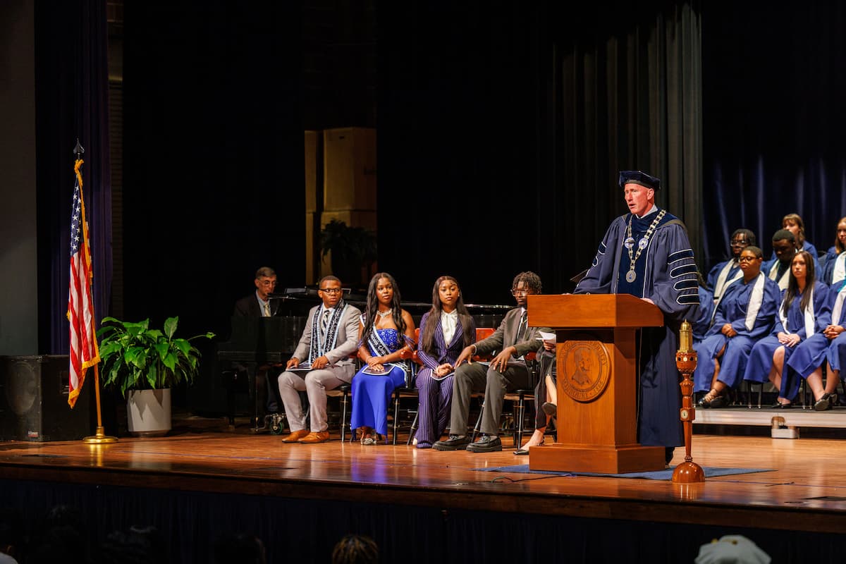 Lincoln University President Moseley stands in academic regalia on a stage behind a podium while giving a speech.