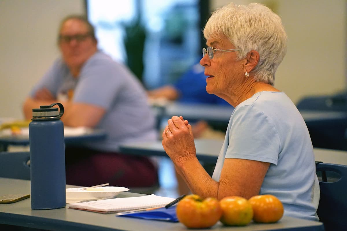 Mary-Jane McCoy, wearing glasses and a light blue shirt, gestures while speaking at a table during the workshop.