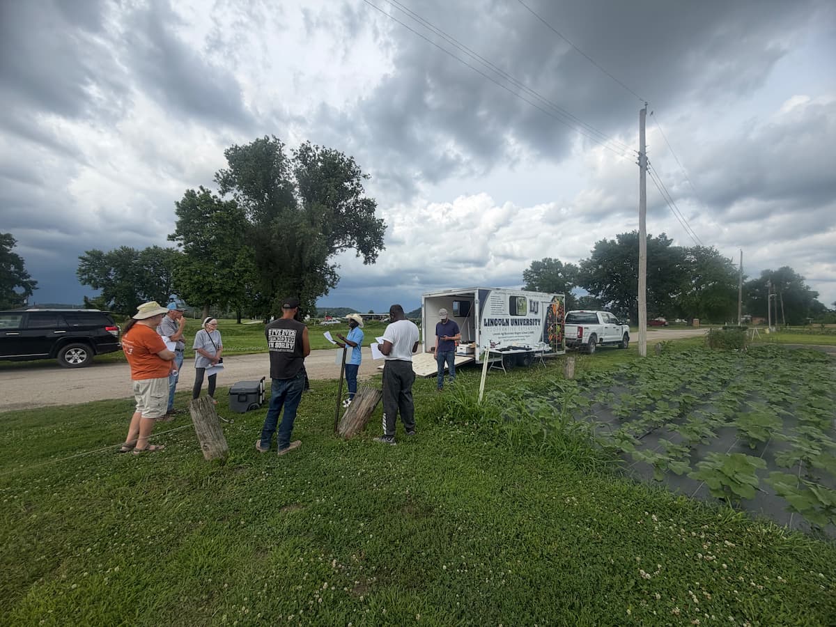 A small group gathered beside a field, signboard, and Lincoln University branded trailer during an event.