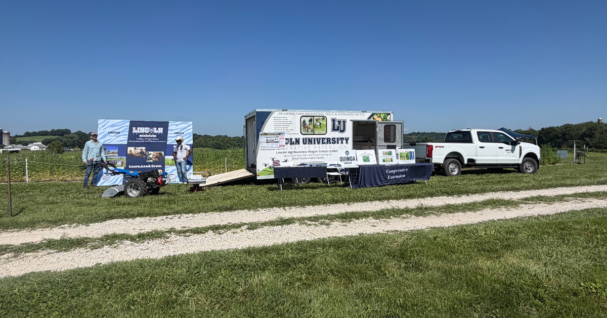 A mobile Lincoln University research trailer, pickup truck, equipment, and two people parked beside a grassy field. 