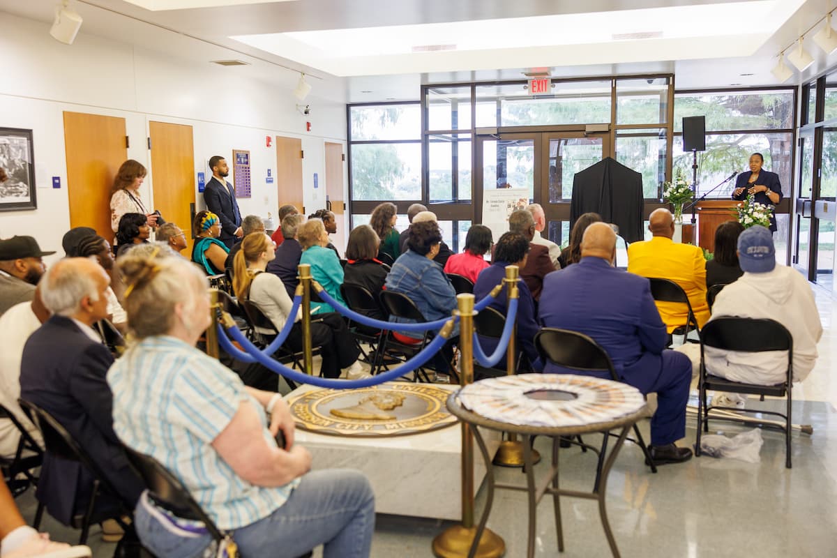 A group of people sit in chairs while listening to speakers at the Dawkins art exhbit.