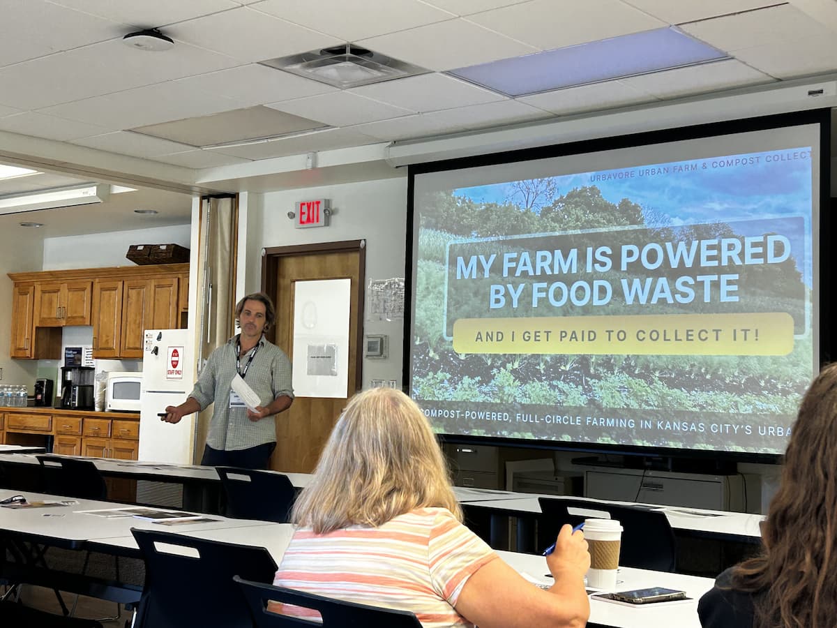 A classroom with people sitting at tables around a screen. On the screen is a slideshow about compost-powered urban farming in Kansas City. 