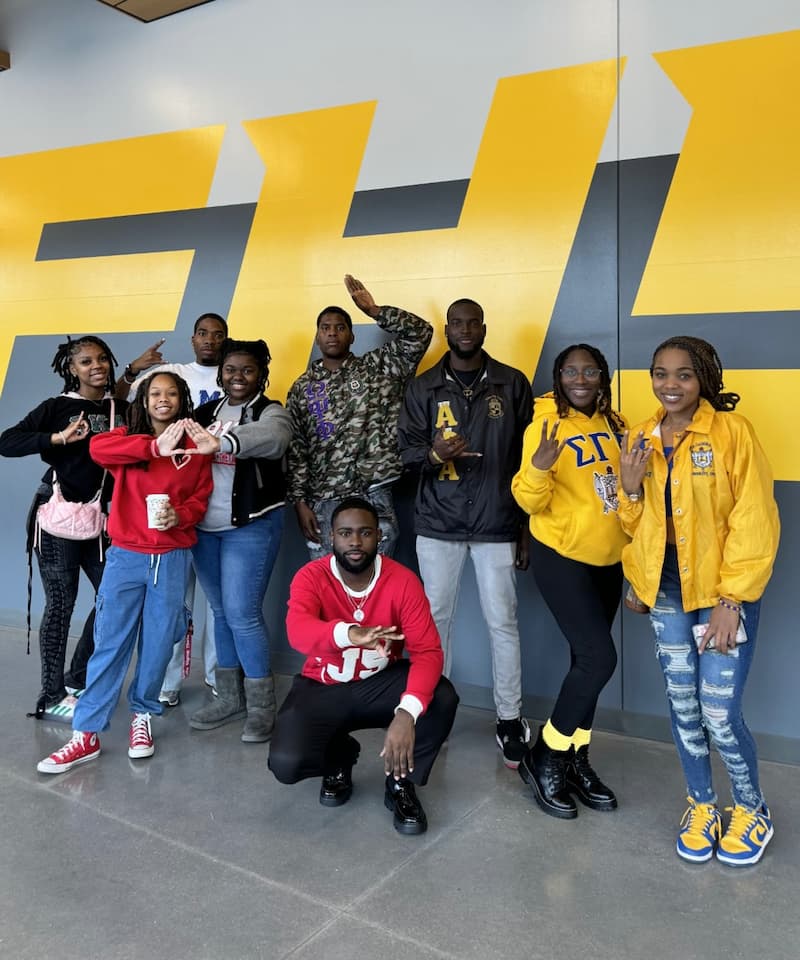 Lincoln University of Missouri members of fraternity and sorority life stand in a clump smiling for a photo while showing off fraternity and sorority signs. 