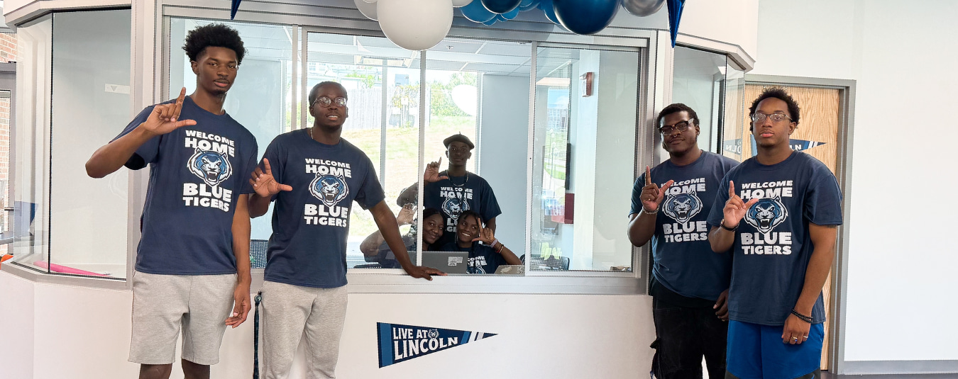 Students wearing “Welcome Home Blue Tigers” shirts pose in a residence hall lobby decorated with blue and white balloons.